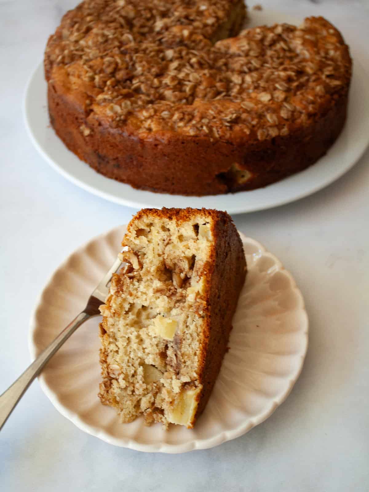 A slice of apple crumb cake on a plate with a fork and the cake behind it.