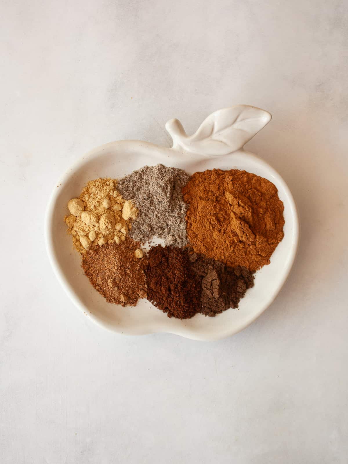Make easy homemade apple pie spice with cinnamon, nutmeg, ginger, allspice, cloves, and cardamom. Perfect for fall baking! The spices for apple pie spice mix including ginger, cardamom, cloves, allspice, nutmeg, and cinnamon on a plate.