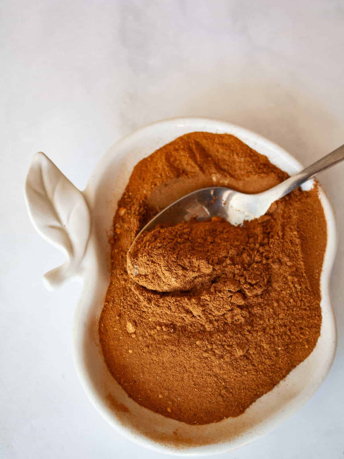 Make easy homemade apple pie spice with cinnamon, nutmeg, ginger, allspice, cloves, and cardamom. Perfect for fall baking! A spoon dips into a bowl in an apple shape that holds apple pie spice.