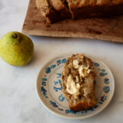 A slice of banana pear with butter on a plate, a loaf on a cutting board, and a pear.