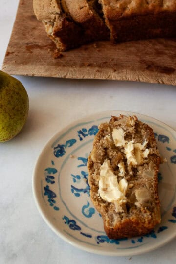 A slice of banana pear with butter on a plate, a loaf on a cutting board, and a pear.