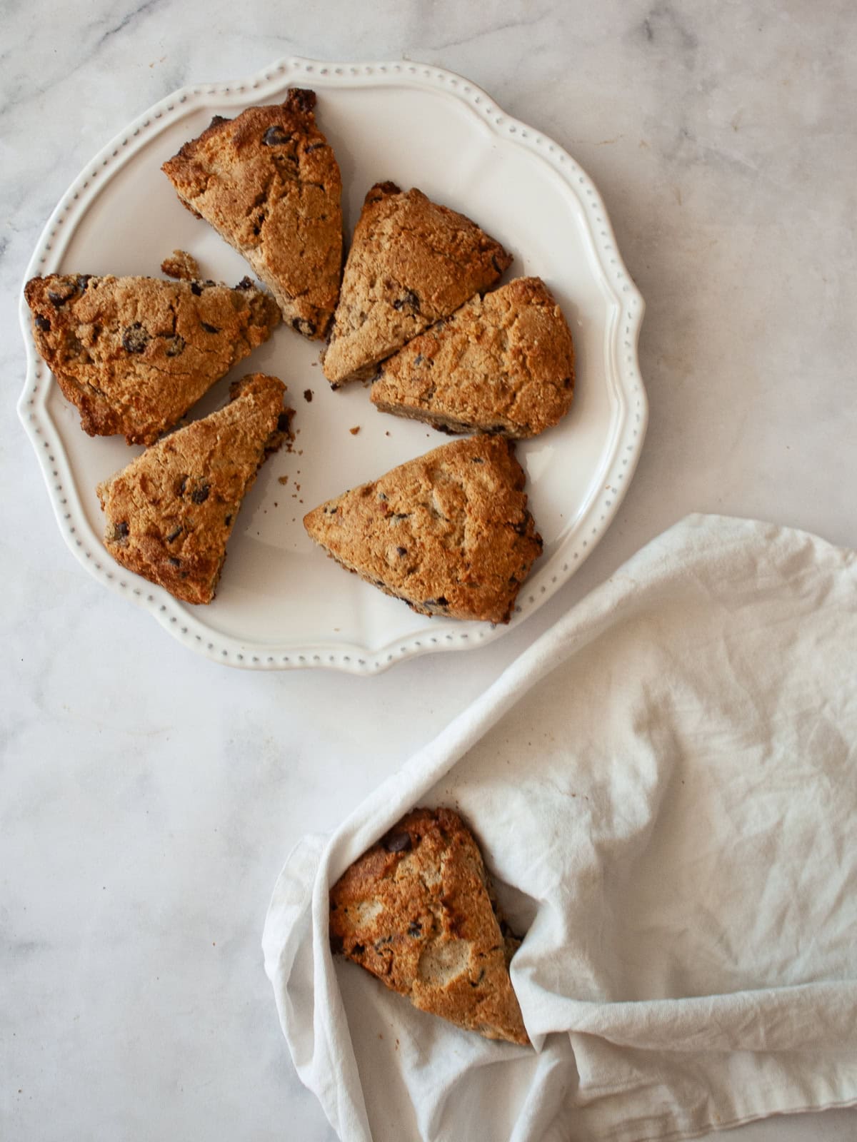 These whole grain banana scones are filled with chocolate chips, cinnamon, and a whole ripe banana for a tender, lightly sweet treat. Banana scones are served on a plate, with a banana scone on a white napkin below it.