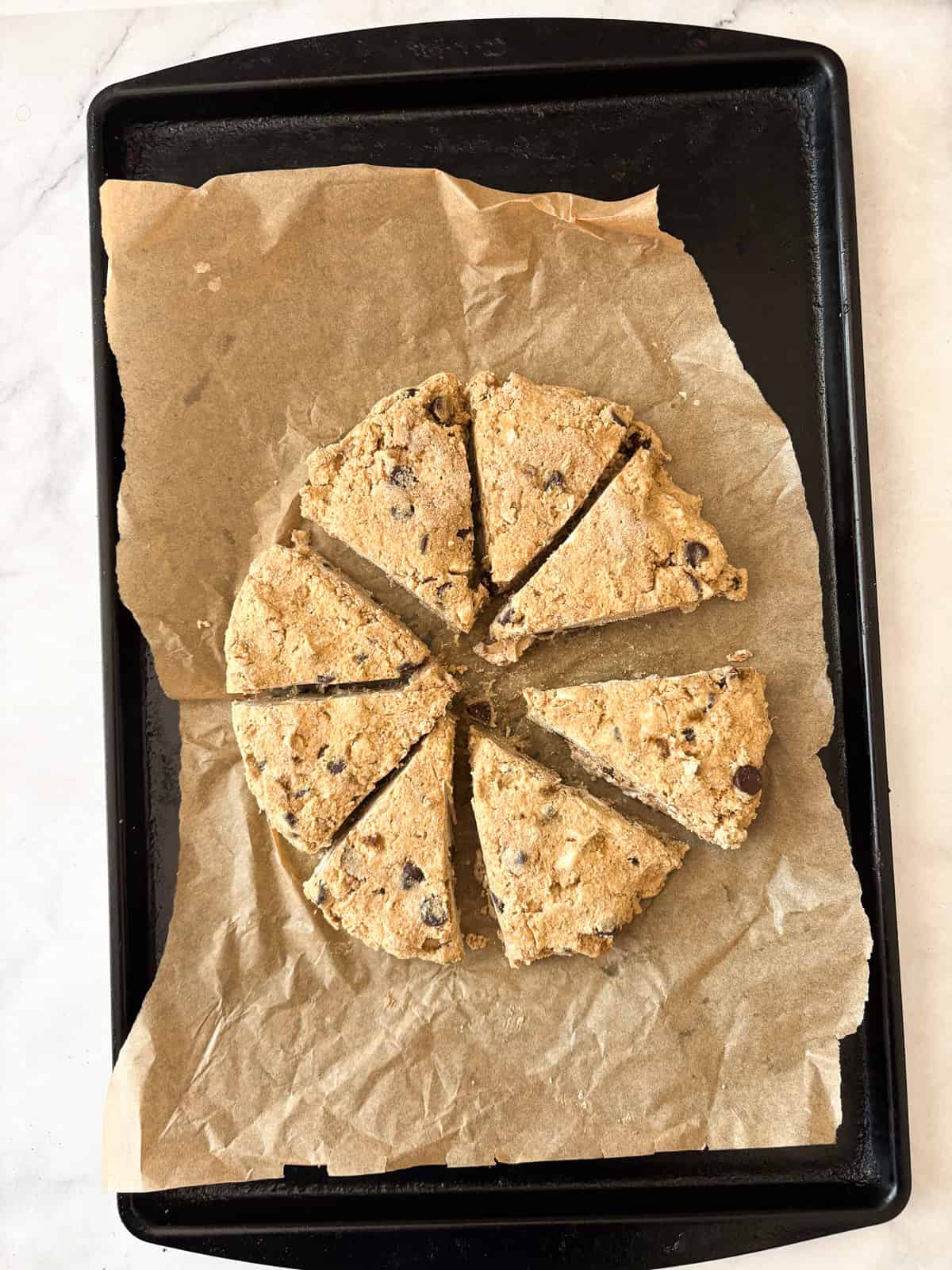 These whole grain banana scones are filled with chocolate chips, cinnamon, and a whole ripe banana for a tender, lightly sweet treat. Banana scones are cut into pieces on a baking sheet.