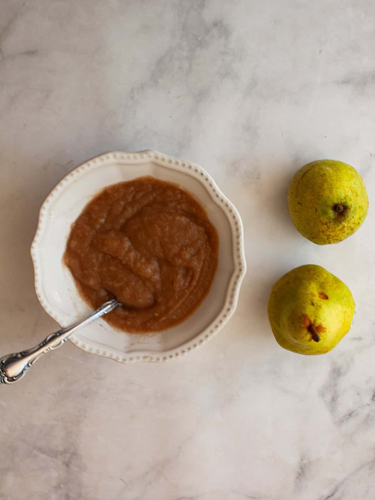 A smooth, sweet pear sauce that's best made with ripe pears! Add cinnamon and maple syrup if you like, or enjoy it plain. A bowl of pear sauce with a serving spoon and two green pears.