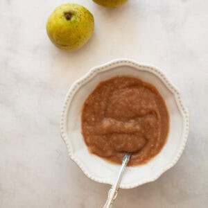 Two pears next to a bowl of pear sauce with a spoon.