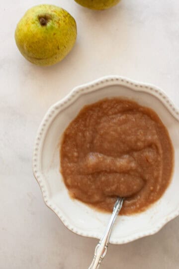 Two pears next to a bowl of pear sauce with a spoon.