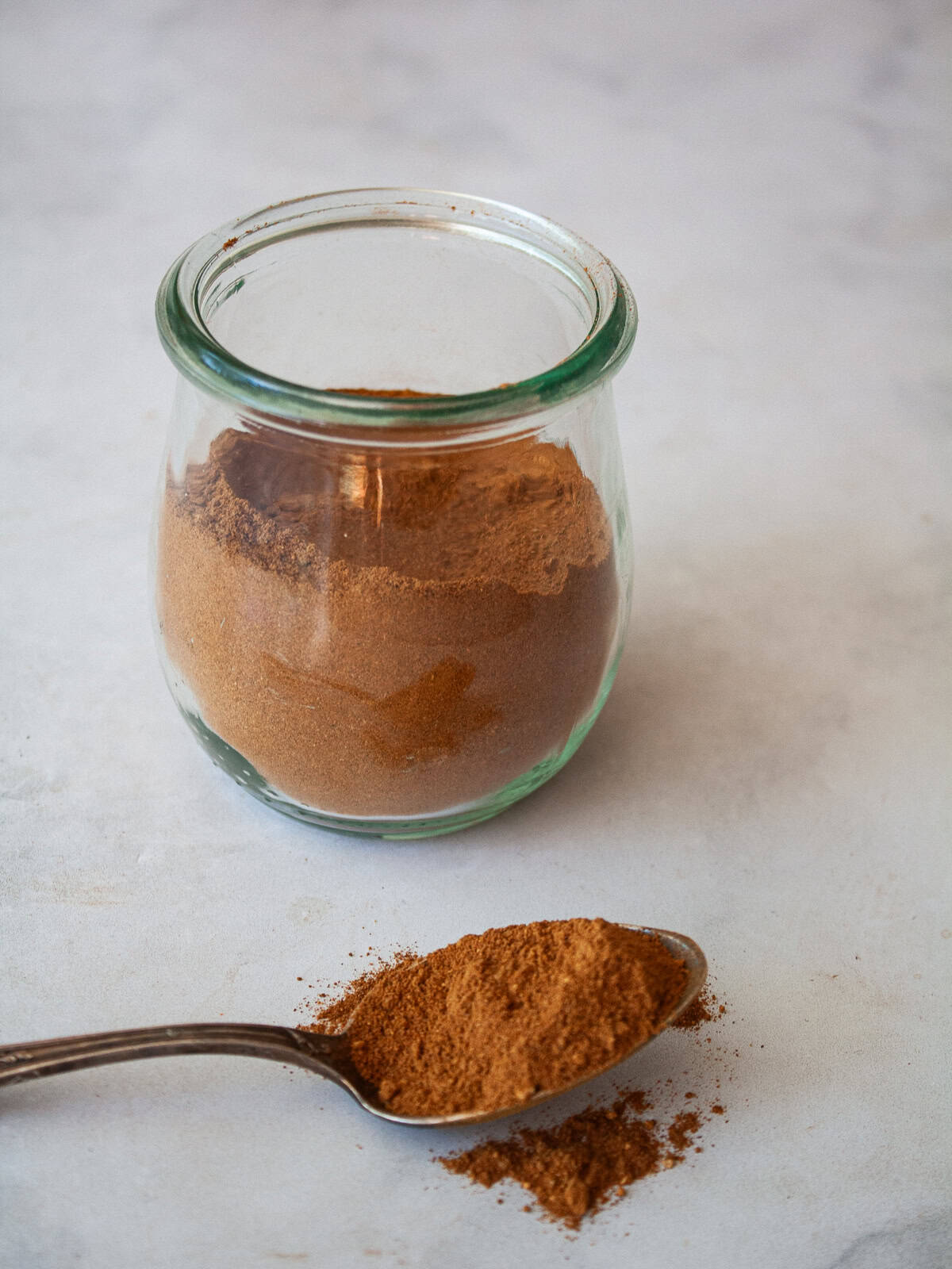 A homemade, versatile pumpkin spice blend with cinnamon, nutmeg, ginger, cloves, and allspice. Great in pies, muffins, quick breads, and more! A spoon with homemade pumpkin spice blend in front of a jar of spice blend.
