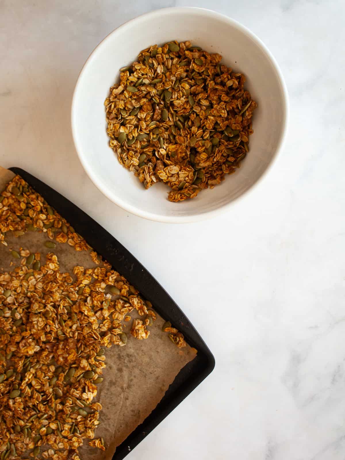 Enjoy this pumpkin spice granola that's made with pumpkin puree, warm spices, crunchy walnuts and pumpkin seeds, olive oil, and maple syrup. A white bowl of pumpkin spice granola next to a baking sheet of granola.